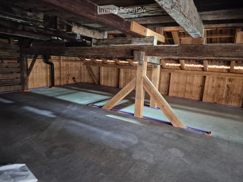 Empty attic space with exposed wooden beams and concrete floor, featuring wooden crates and storage boxes along the walls.
