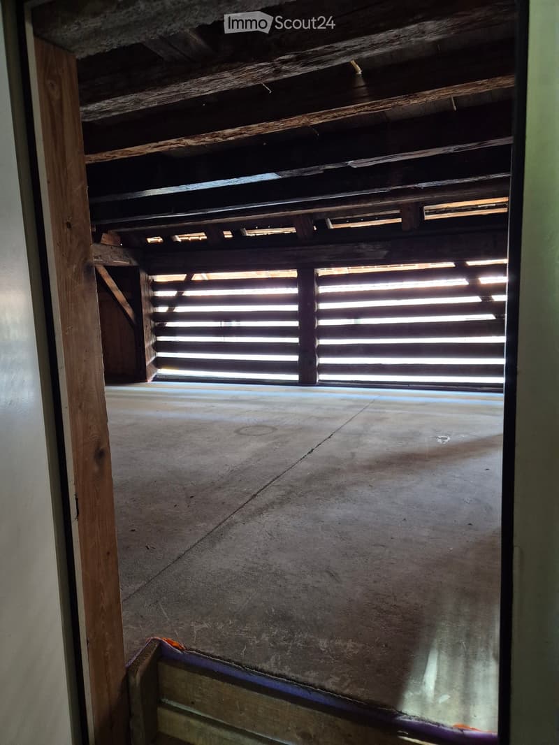 Empty room with concrete floor, wooden ceiling beams, and a window with horizontal slats allowing natural light to enter.
