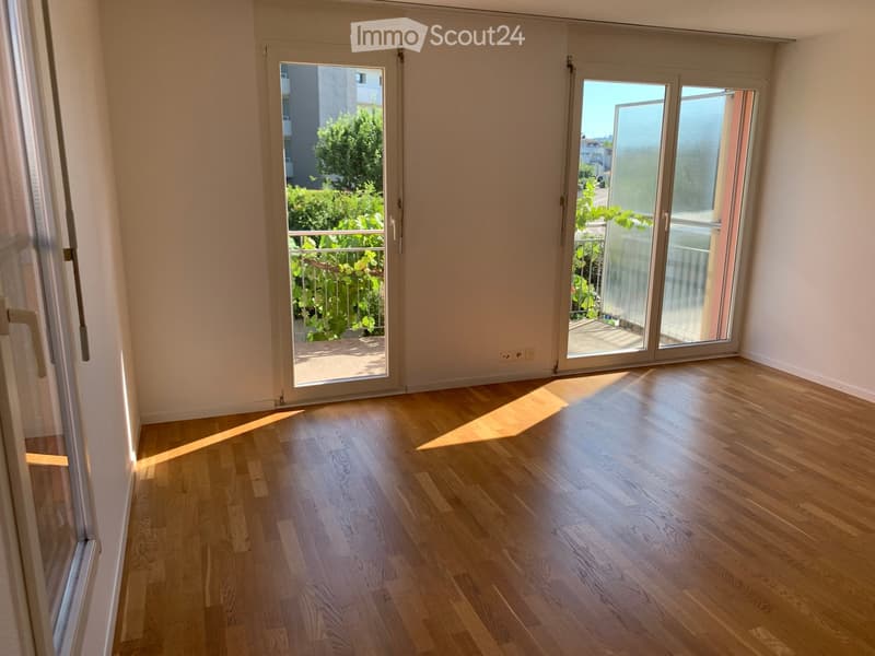 Empty room with wooden flooring and large sliding glass doors leading to a balcony with greenery visible outside.
