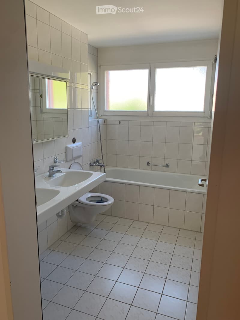 Modern bathroom with white tiles, double sink, toilet, bathtub, and large windows providing natural light.
