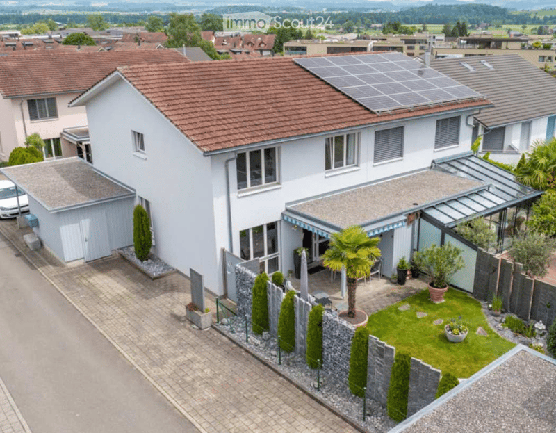 House. A two-story white house with a red-tiled roof, solar panels on the roof, and a modern garage. The house features large windows and a well-maintained garden with a palm tree, shrubs, and a small lawn.