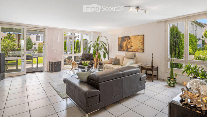 Living Room. Bright living room with large windows and a view of the garden. Features a black leather sofa, a beige armchair, a coffee table, a floor lamp, and decorative items including a picture of a lion and a vase with flowers.