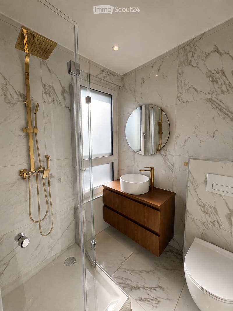 Modern bathroom with marble tiles, a glass-enclosed shower, a wooden vanity with a sink, a round mirror, and a toilet.