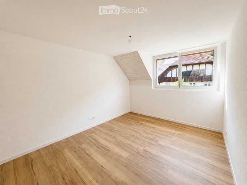 Empty room with light wooden flooring, white walls, and a large window showing a view of a neighboring building with a red-tiled roof.