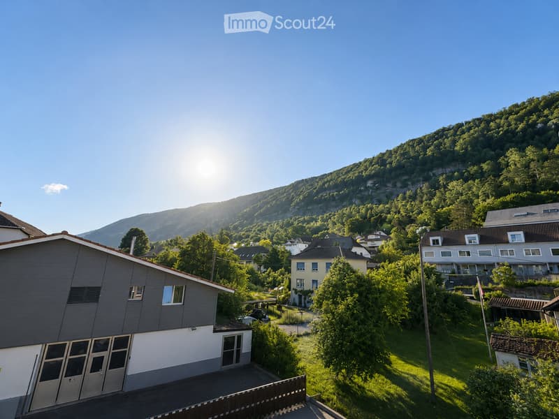 Residential area with multiple houses, surrounded by greenery and a forested hill in the background.