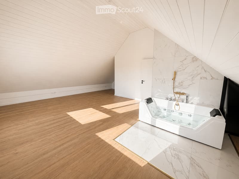 Modern bathroom with wooden flooring, white walls, and a sleek bathtub with a glass enclosure. The space features a marble accent wall and a sloped ceiling with exposed wooden beams.