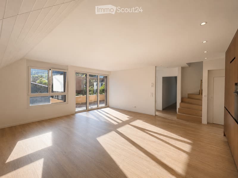 Bright, empty room with wooden flooring, large windows, and sliding glass doors leading to a balcony. The ceiling has a modern, slanted design with recessed lighting.