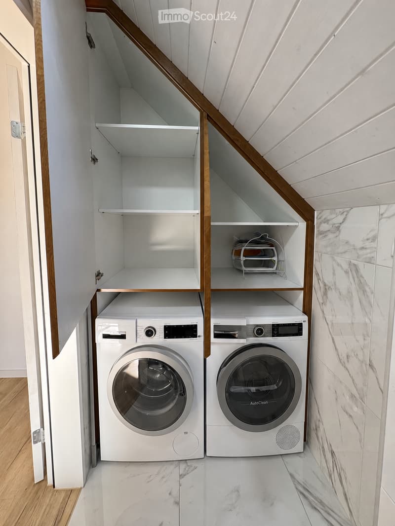 Modern laundry area with white washing machine and dryer, marble flooring, built-in white cabinets with wooden trim, and a small shelf holding a charging station.