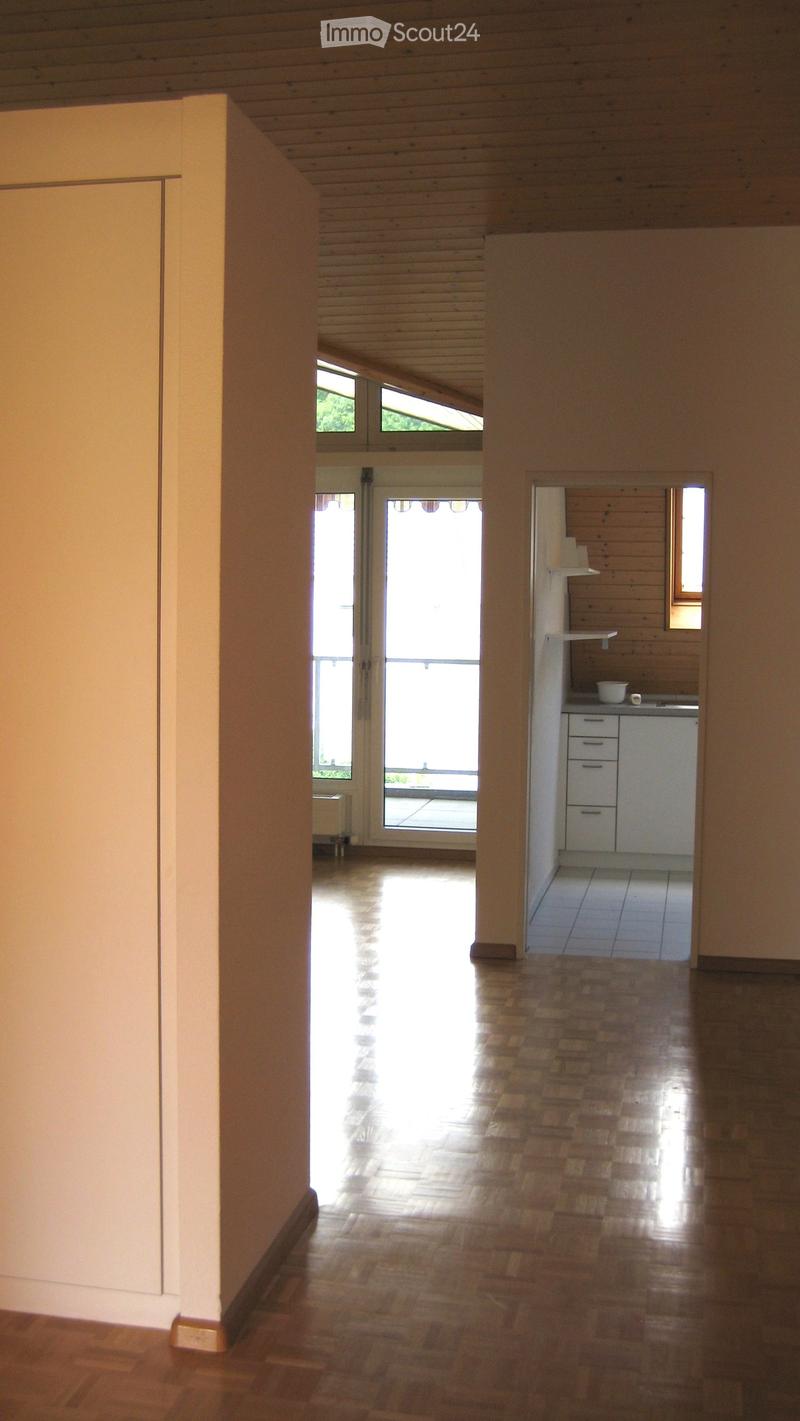 Bright, open hallway with wooden flooring and white walls, leading to a small kitchen area with white cabinets and a sink.