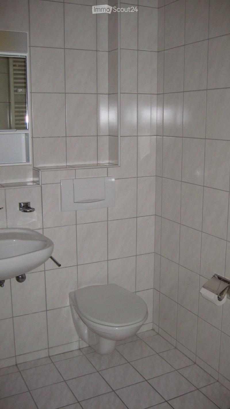 A small bathroom with white tiled walls and floor, featuring a toilet, a sink, a mirror, and a toilet paper holder.