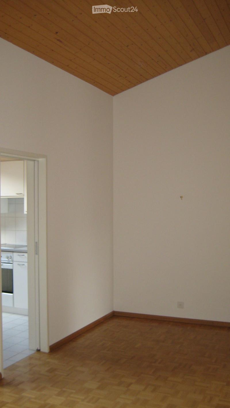 Empty room with light-colored walls and wooden flooring, featuring a slanted wooden ceiling and an open doorway leading to another room.