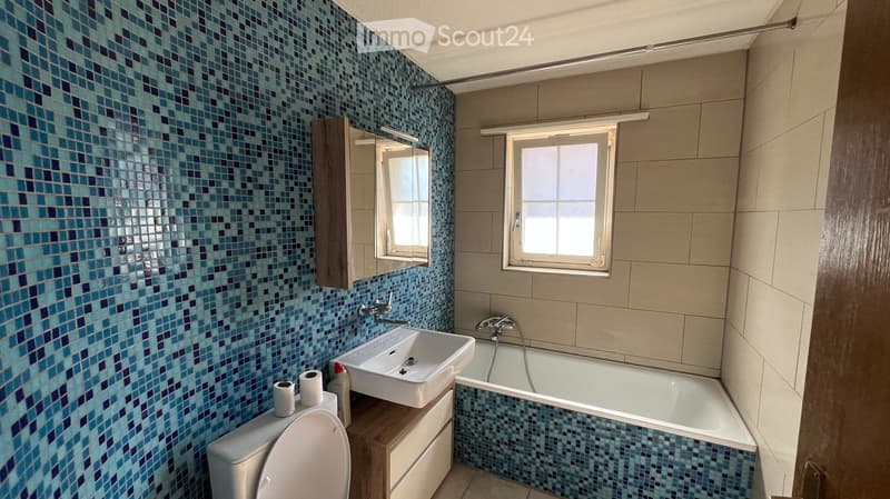 Modern bathroom with blue mosaic tiles, a white sink, a bathtub, and a toilet. Features a large mirror and two windows for natural light.