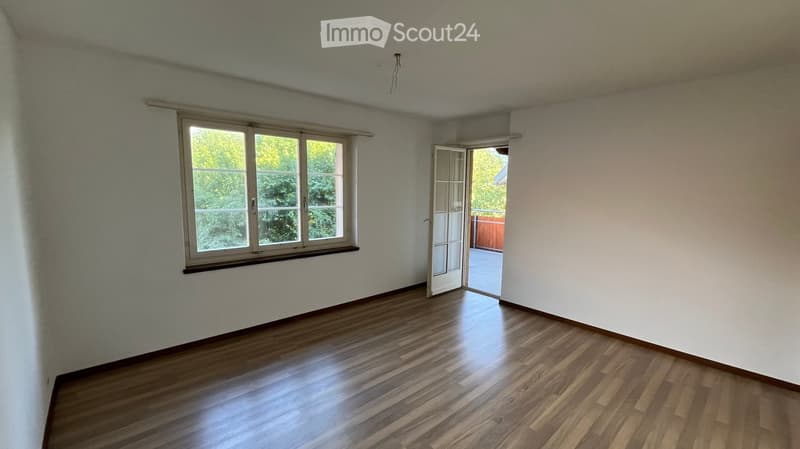 Empty room with wooden flooring, white walls, large window, and open door leading to a balcony or outdoor area.