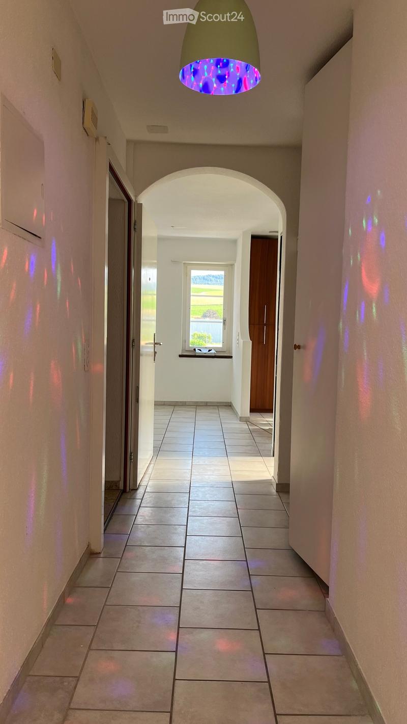 A modern hallway with tiled flooring, white walls, and a colorful hanging light fixture. An arched doorway leads to another room with a window and a small table.