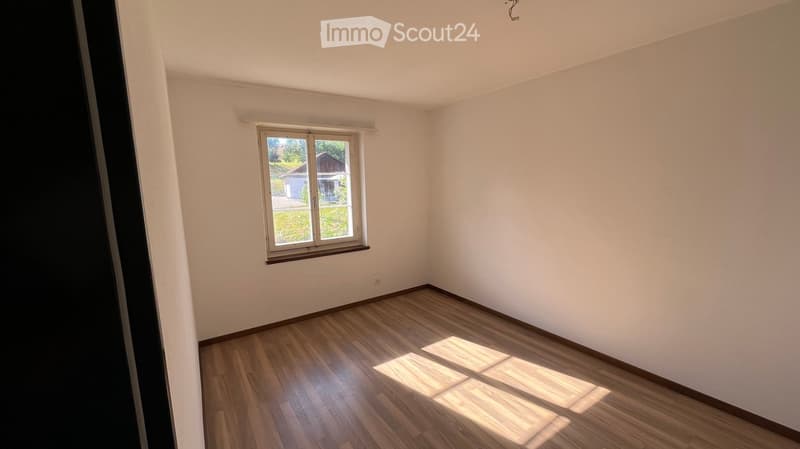 Empty room with wooden flooring and white walls, featuring a single window with a view of a garden and a house outside.