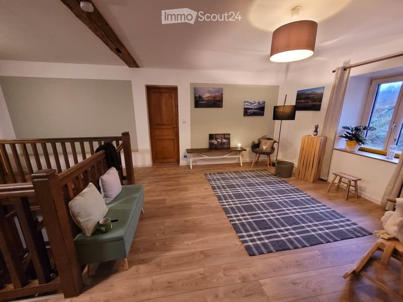 Cozy living room with wooden floors, a green sofa, a bench, a chair, a small table, a floor lamp, a plaid rug, and several picture frames on the wall.