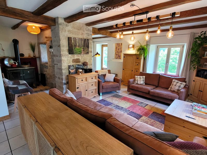Cozy living room with exposed wooden beams, stone fireplace, large windows, and comfortable seating including a sofa, armchair, and ottoman. Wooden coffee table, pendant lights, and decorative items complete the space.
