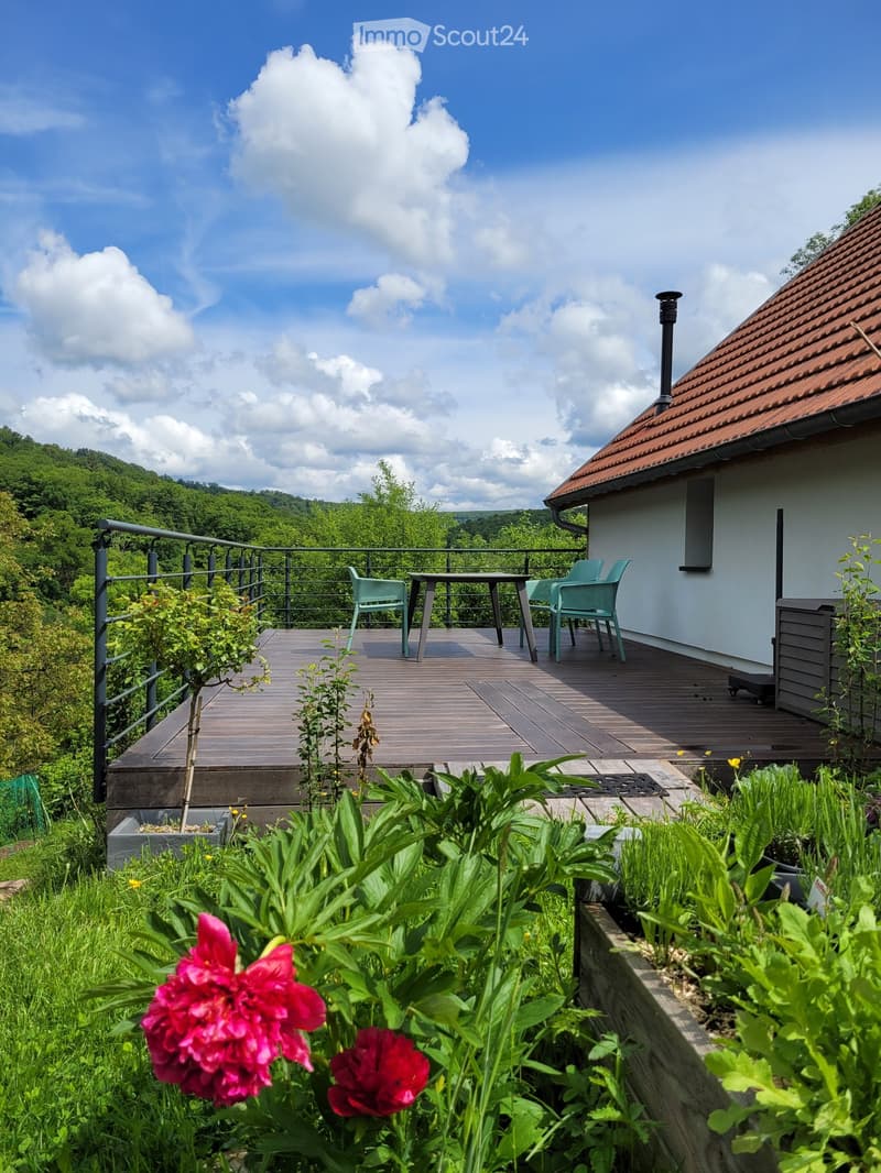 Wooden deck with outdoor furniture, surrounded by greenery and flowers, overlooking a scenic valley.