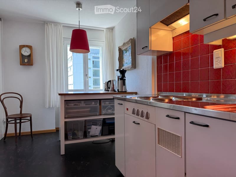 Modern kitchen with white cabinets, stainless steel appliances, a central island, and a pendant lamp. Features a coffee maker, a chair, and a window with white curtains.