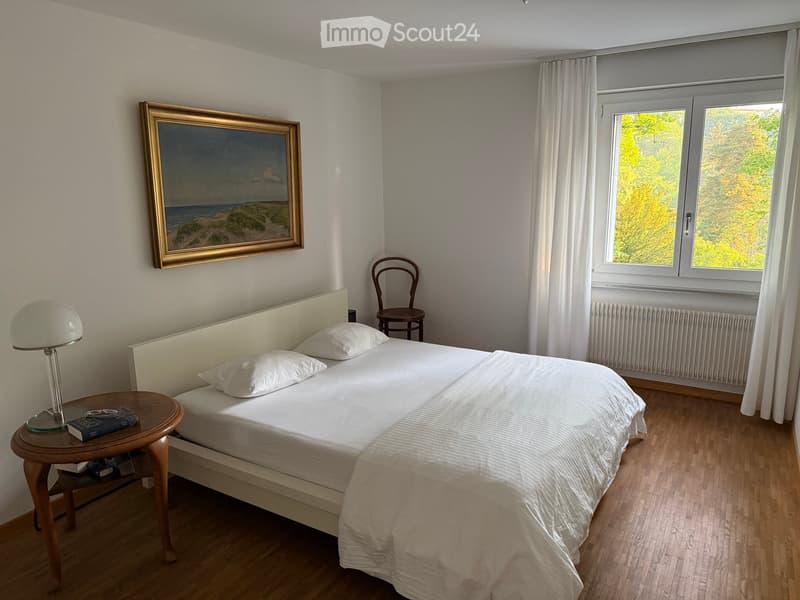 A well-lit bedroom with a white bed, nightstand, lamp, chair, and a large painting on the wall. The room has wooden flooring and large windows with white curtains.