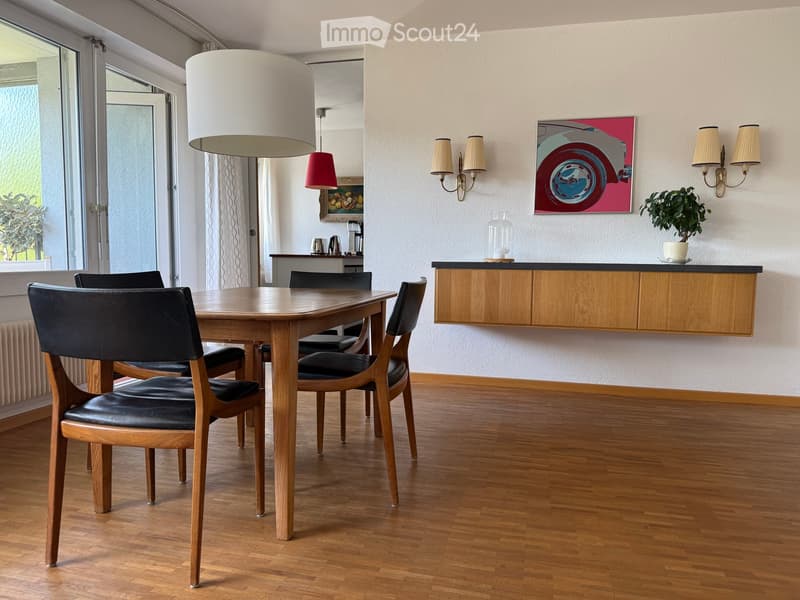 Dining room with a wooden dining table and four black leather chairs, a wooden cabinet, pendant light, wall-mounted lamps, a painting, and a small plant on the counter.