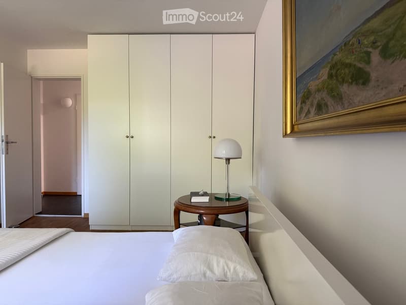 Modern bedroom with a white bed, round wooden nightstand, table lamp, large white wardrobe, and a framed painting on the wall.