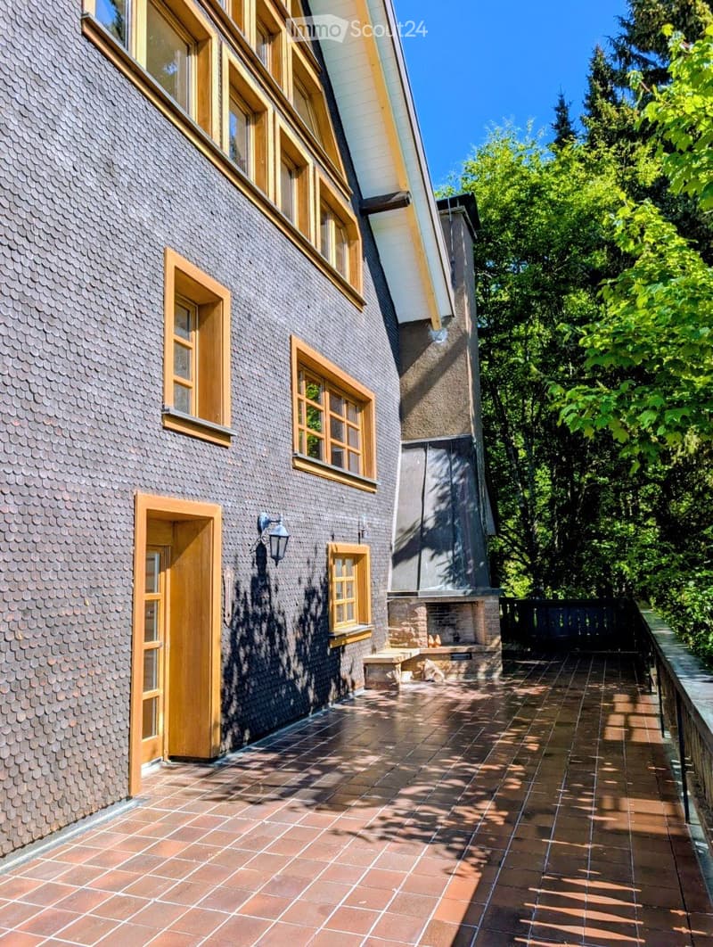 Two-story house with dark grey tiled exterior, wooden-framed windows, and a covered entrance leading to a tiled patio area surrounded by greenery.
