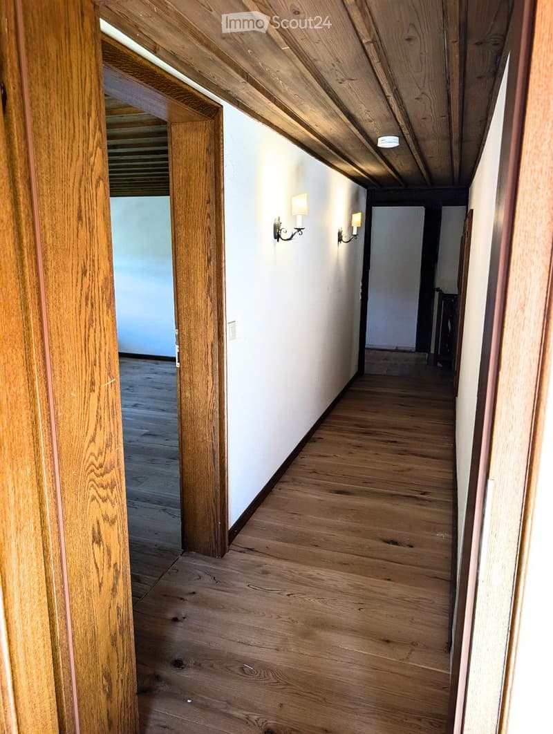 Wooden-floored hallway with white walls, wooden ceiling beams, and wall-mounted light fixtures. The hallway leads to an open doorway on the left and a closed door on the right.