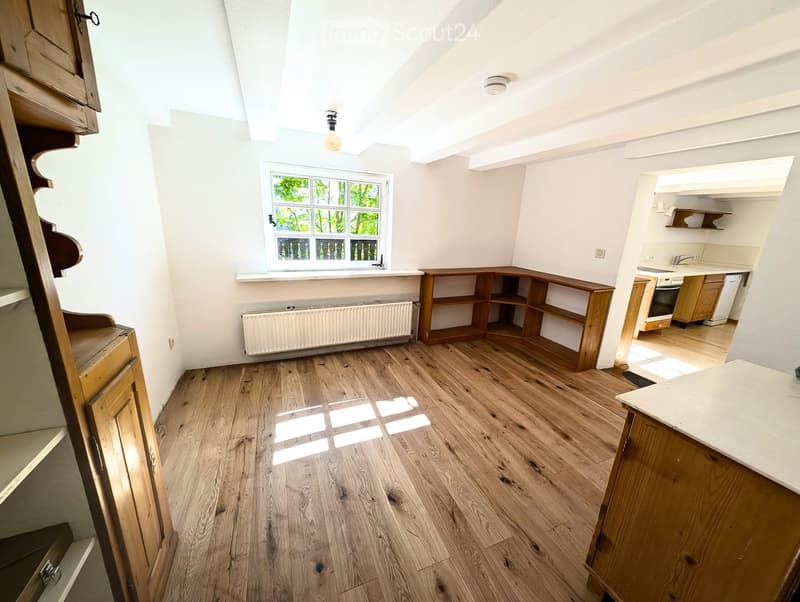 Empty room with wooden flooring, white walls, and a window with multiple panes. There is a radiator below the window and a wooden shelf unit against one wall. The room is connected to a kitchen area with cabinets and appliances visible in the background.