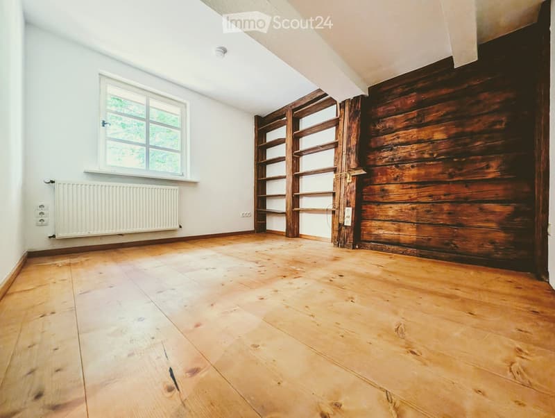 Empty room with wooden flooring, white walls, a window, a radiator, and a wooden shelving unit.