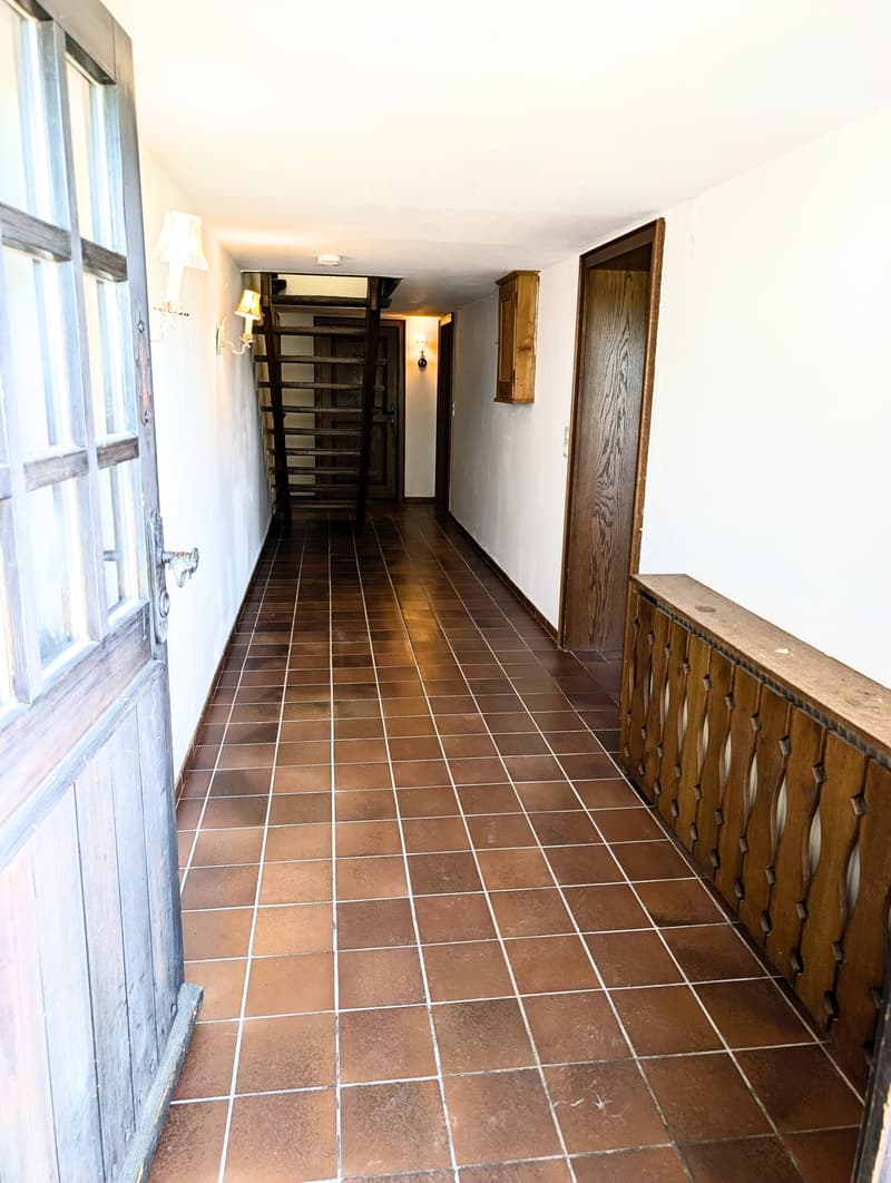 A long hallway with brown tiled flooring, white walls, wooden doors, and a staircase leading upwards. The hallway features wall-mounted lamps and a wooden railing on one side.