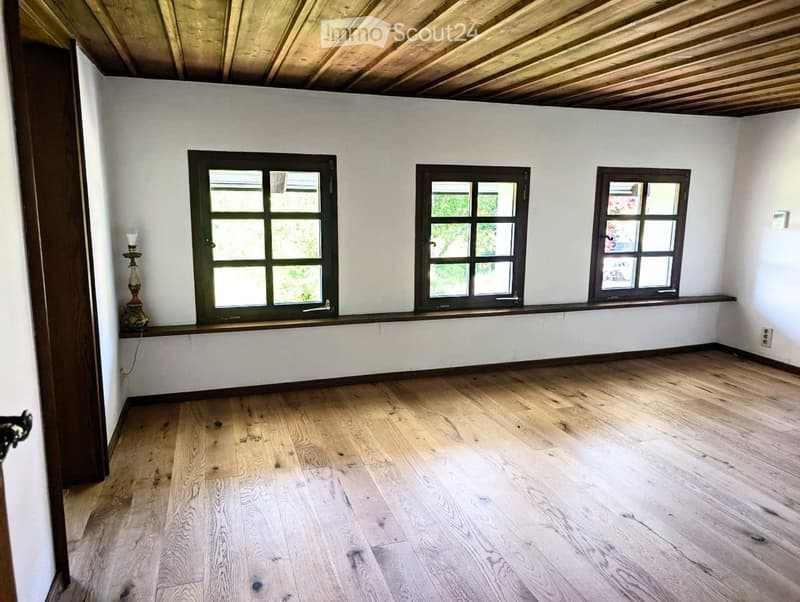 Empty room with wooden flooring, white walls, and three windows with dark frames. A small decorative lamp is placed on a windowsill.