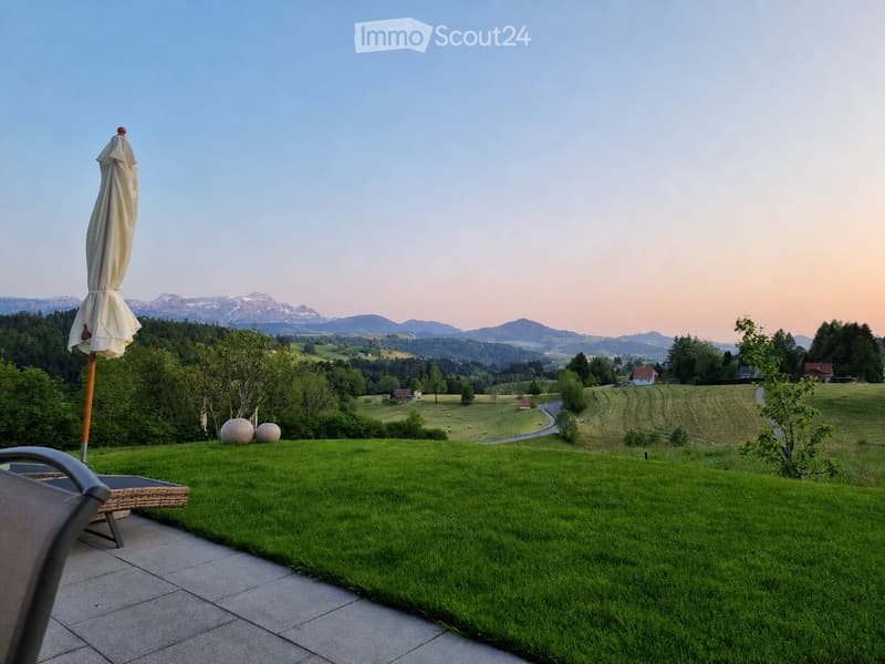 A serene garden with a well-maintained lawn, a closed umbrella, and a scenic view of rolling hills and mountains in the background.