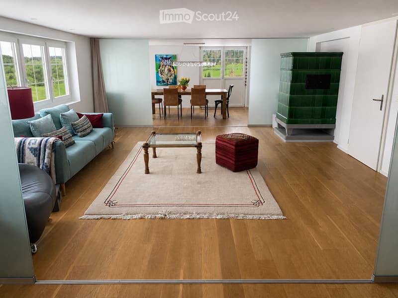 Bright living room with wooden flooring, large windows, a blue sofa, coffee table, ottoman, dining area with chairs and table, and a green fireplace.