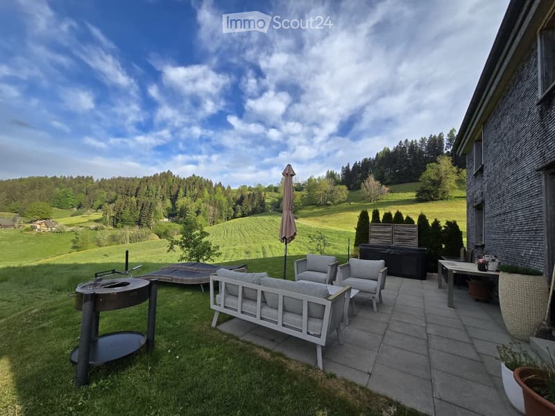 Outdoor patio with a sofa, chairs, a table, umbrella, grill, and fire pit, overlooking a scenic countryside view.
