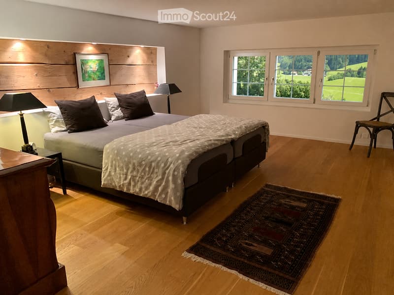 Cozy bedroom with wooden flooring, large bed with dark bedding, nightstand with lamp, chair, and a patterned rug.