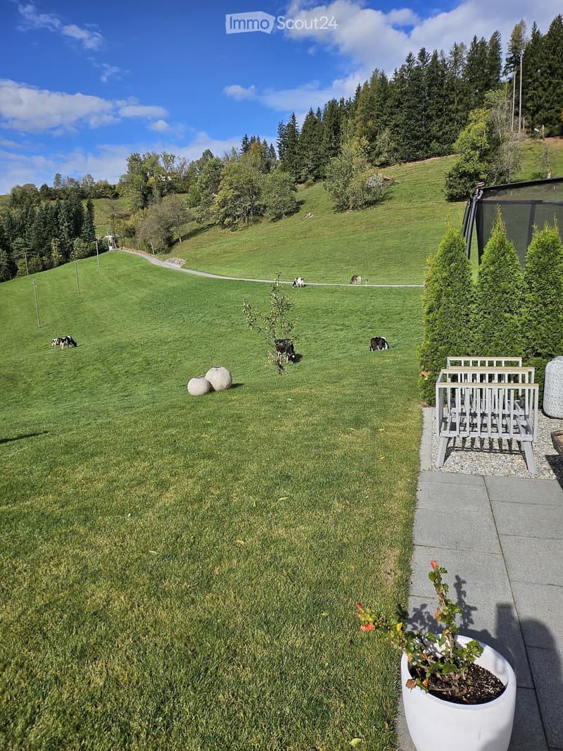 A well-maintained garden with a grassy lawn, grazing cows in the background, and a small seating area with a white planter and decorative spheres.