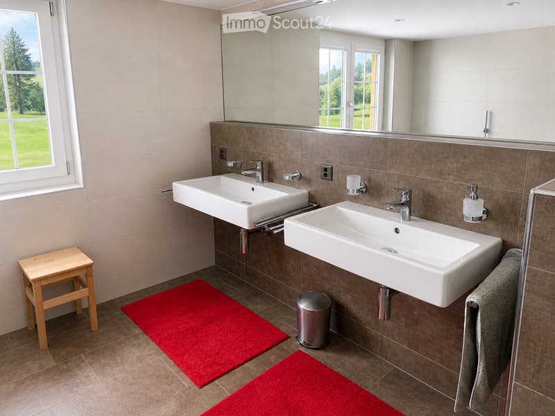 Modern bathroom with two white sinks, chrome faucets, a large mirror, a wooden stool, red rugs, and a towel on a rack.