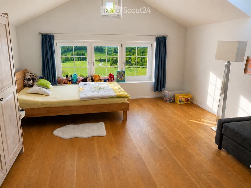 Bright bedroom with wooden flooring, large windows overlooking a green landscape, a bed with pillows and a blanket, a sofa, a wardrobe, a floor lamp, and a toy box.
