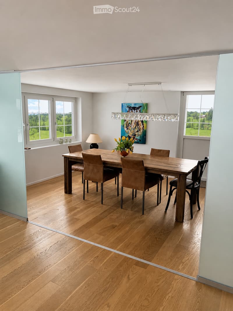 Dining room with wooden floor, large wooden dining table surrounded by brown leather chairs, a table lamp, a flower vase with yellow tulips, a painting on the wall, and a modern chandelier hanging from the ceiling. Large windows offer a view of a green la