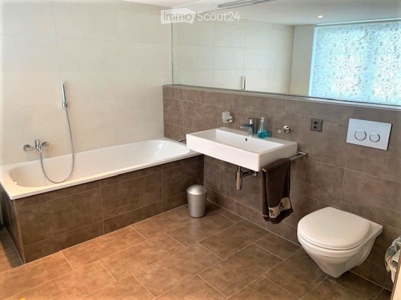 Modern bathroom with a bathtub, sink, toilet, large mirror, and a towel hanging on a rack. The walls are tiled in a neutral color scheme, and the floor is covered with brown tiles.