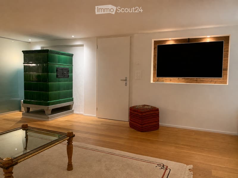 Modern living room with wooden flooring, a glass coffee table, a red ottoman, and a mounted television on the wall. A green tiled fireplace is visible in the background.