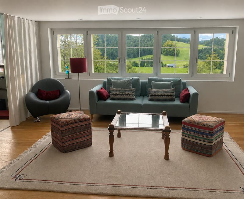 Bright living room with large windows offering a scenic view, featuring a teal sofa, dark armchair, patterned ottomans, a glass coffee table, and a floor lamp.