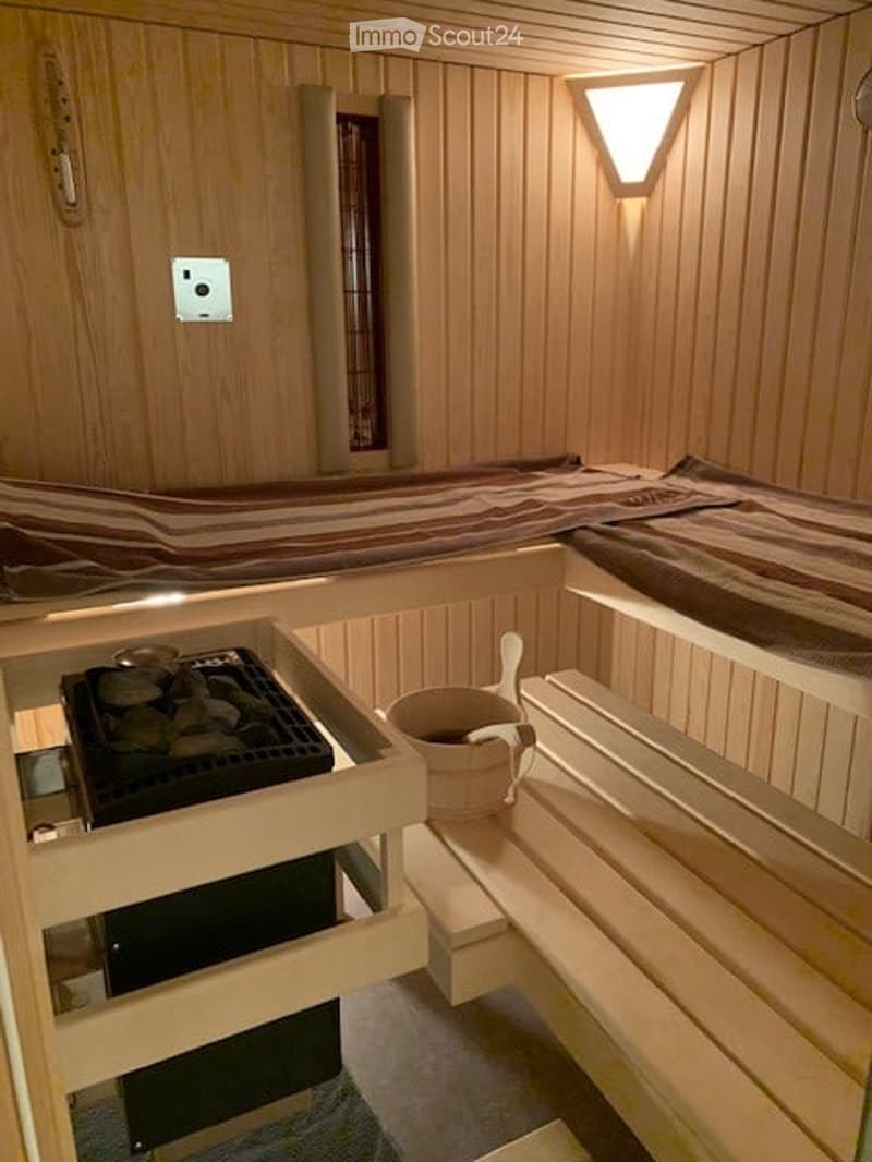 A small, cozy sauna room with wooden walls and ceiling, featuring a wooden bench, a wooden bucket, a wooden stove, and a bed with striped bedding.