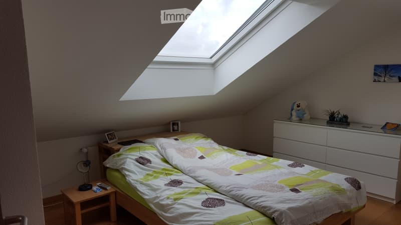 Cozy attic bedroom with a skylight window, a double bed with a colorful duvet, a wooden nightstand with a fan and remote controls, a white dresser with a plant and a stuffed toy, and framed pictures on the walls.
