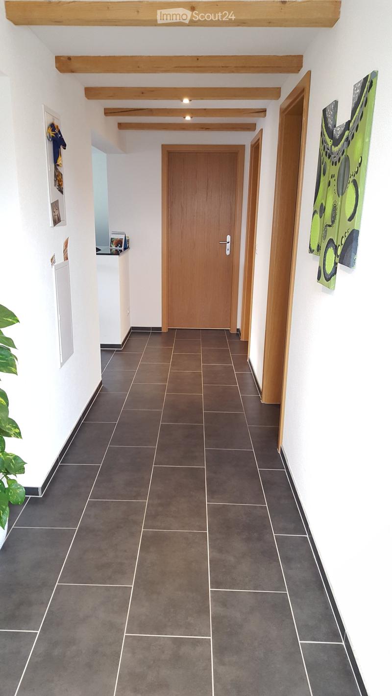 Clean hallway with dark tiled flooring, white walls, wooden doors, and exposed wooden beams on the ceiling. Features a small shelf with books and a plant on the left side.