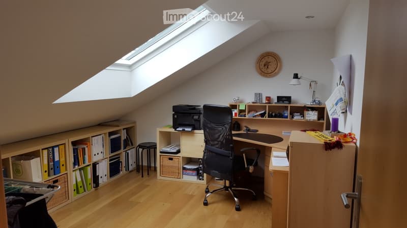 Cozy attic office with wooden flooring, slanted ceilings, and a skylight. Features a desk with a chair, printer, bookshelf, and a small stool.