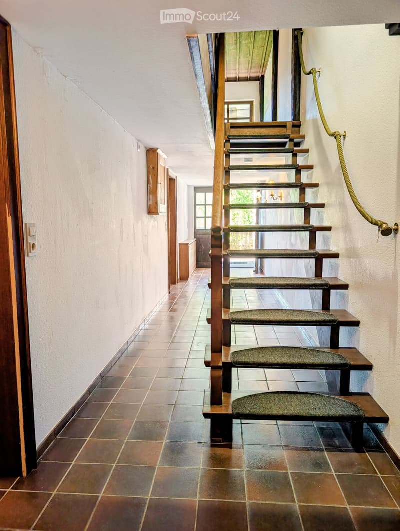 A narrow hallway with tiled flooring, wooden doors, and a staircase with wooden railings and green carpeted steps leading upwards.