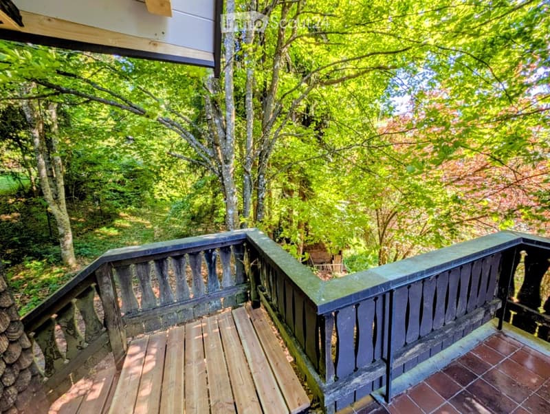 Wooden balcony with a dark railing, surrounded by lush green trees and foliage, with a wooden deck and tiled floor.