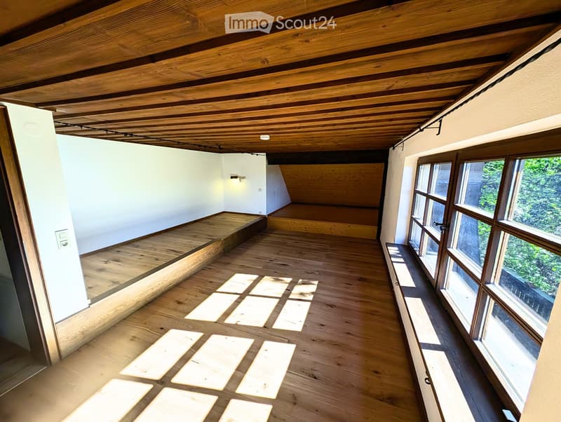 An empty room with wooden flooring and exposed wooden ceiling beams. Large windows on one side allow natural light to fill the space. The room features built-in wooden benches along the walls and a raised platform area in the corner.